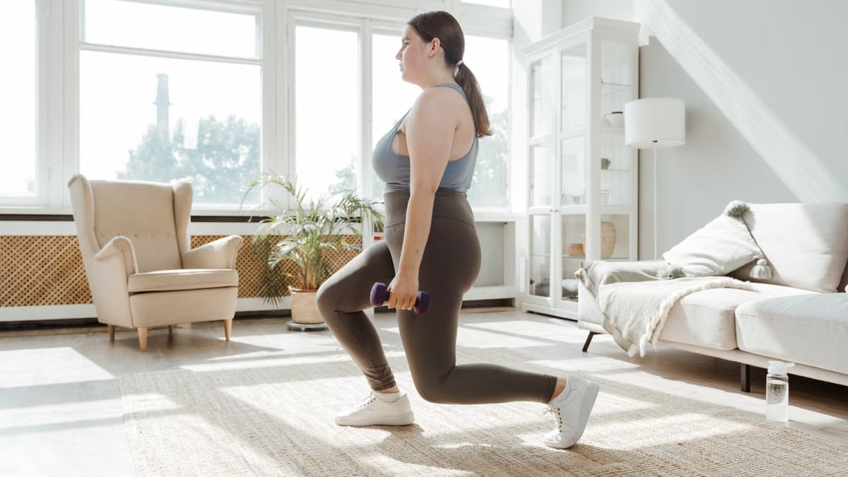 A woman in activewear doing lunges with dumbbells in a bright living room.