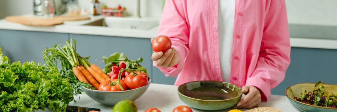Woman in pink overshirt holds tomato in kitchen with fresh fruits and veggies.